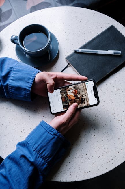 A person views Instagram on a smartphone at a cafe table with coffee, notebook, and pen.